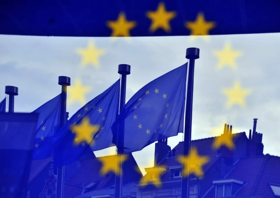 Imaginea prezintă mai multe steaguri ale Uniunii Europene fluturând, cu o suprapunere a stelelor UE, pe un fundal de cer și clădiri.