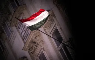 O steag cu dungi orizontale roșii, albe și verzi este atașat unui stâlp de pe fațada unei clădiri de piatră sub un cer întunecat.