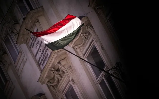 O steag cu dungi orizontale roșii, albe și verzi este atașat unui stâlp de pe fațada unei clădiri de piatră sub un cer întunecat.