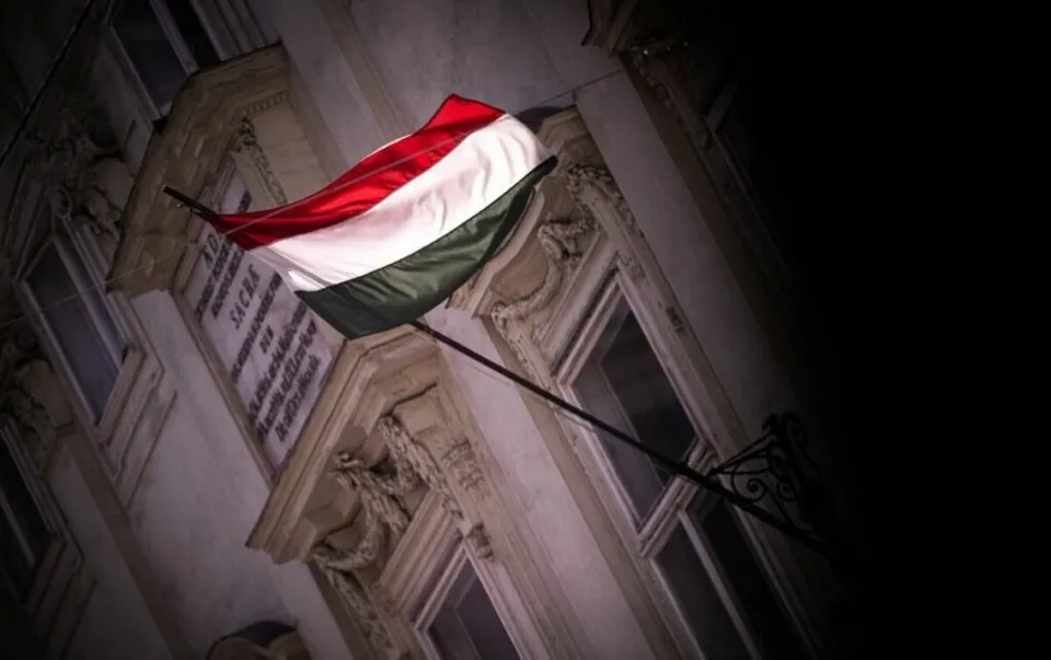 O steag cu dungi orizontale roșii, albe și verzi este atașat unui stâlp de pe fațada unei clădiri de piatră sub un cer întunecat.