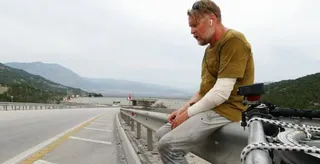 Un bărbat cu părul scurt și barba căruntă stă așezat pe o barieră metalică de protecție de lângă un drum asfaltat.