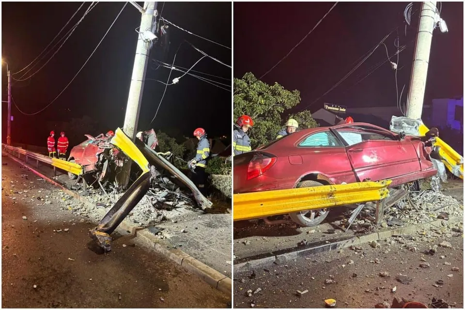 Un autoturism roșu este lovit de un stâlp de beton lângă o barieră metalică galbenă, în timp ce persoane cu echipament reflectorizant stau în apropiere.