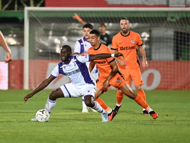 Cinci jucători de fotbal sunt pe un teren de gazon verde, în timp ce unul în echipament alb și violet dispută mingea cu doi jucători în echipament portocaliu.