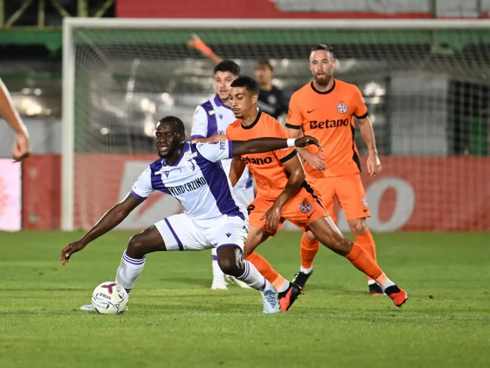 Cinci jucători de fotbal sunt pe un teren de gazon verde, în timp ce unul în echipament alb și violet dispută mingea cu doi jucători în echipament portocaliu.