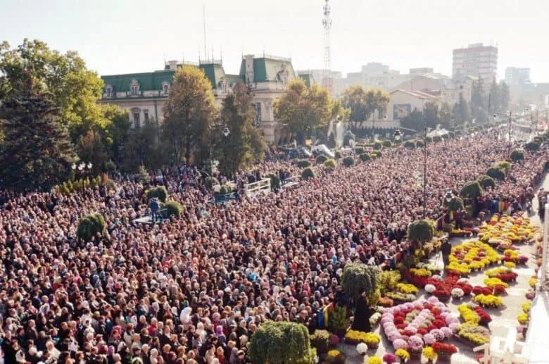 O mulțime mare de oameni este adunată pe o stradă largă lângă clădiri și copaci, lângă straturi de flori colorate.
