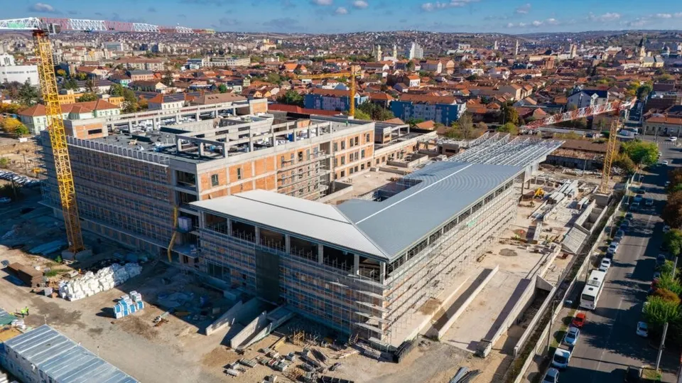 O vedere aeriană prezintă un complex de clădiri în construcție cu schele, macarale și materiale de șantier pe un teren de dimensiuni mari.