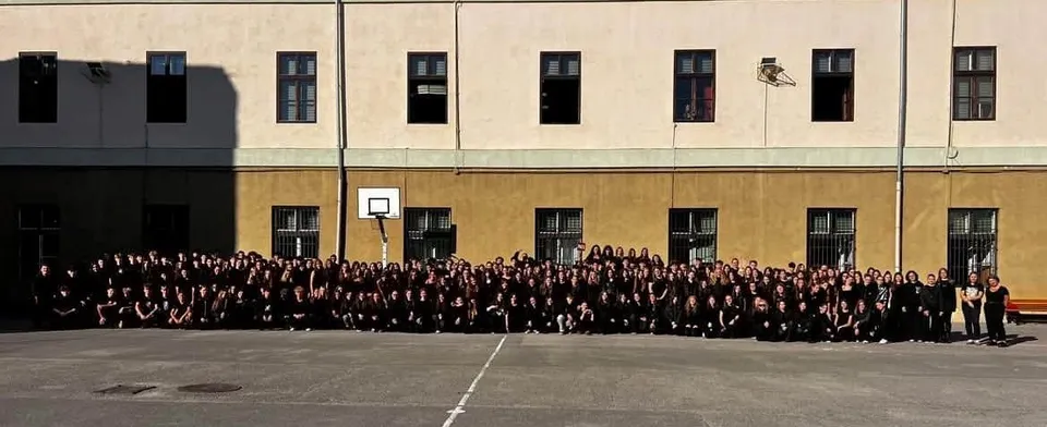 Un grup mare de persoane îmbrăcate în haine negre stă întins pe un teren de sport în fața unei clădiri cu ferestre rectangulare.