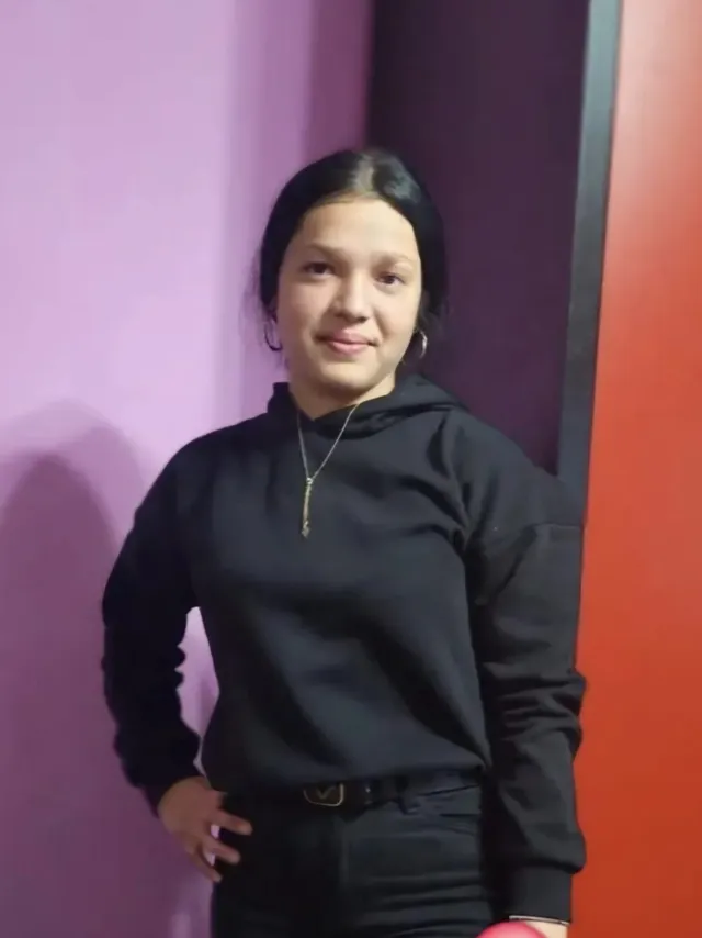 O persoană cu părul negru și o bluză neagră cu glugă stă în picioare în fața unui perete violet și a unuia roșu.
