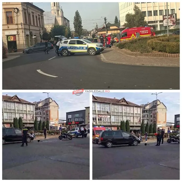 O mașină de poliție albastră și galbenă stă pe un drum lângă un sens giratoriu, în timp ce un vehicul de urgență roșu este parcat în fundal.