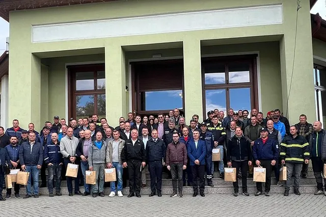 Un grup mare de bărbați stă în rând în fața unei clădiri verzi, mulți dintre ei ținând pungi de carton maro.