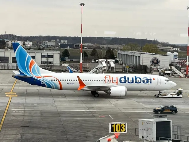 Un avion alb cu însemne FlyDubai este parcat pe o pistă de aeroport lângă diverse vehicule de serviciu și clădiri de terminal.