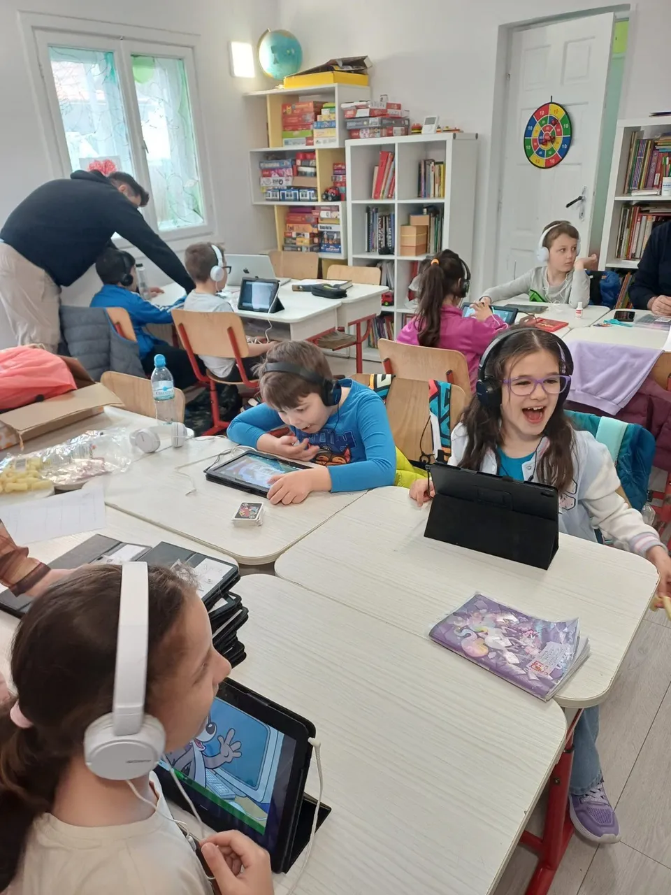 Copii stau la mese albe în interiorul unei încăperi, folosind tablete și căști, în timp ce un adult stă aplecat lângă un copil.