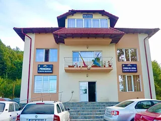 O clădire cu două etaje prezintă ferestre, un balcon cu ghivece de flori și două panouri informative pe fațadă, având mașini parcate în prim-plan.