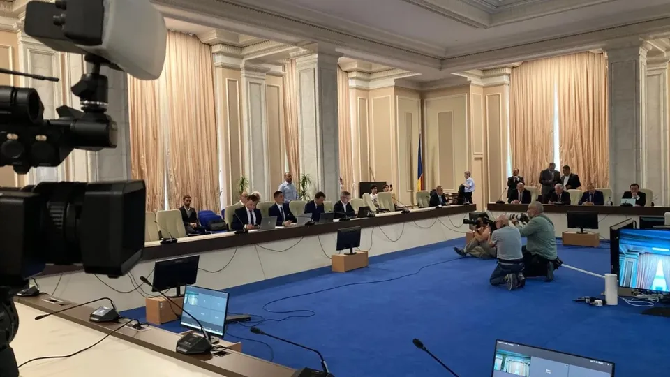 O cameră de filmare este în prim-plan, în timp ce mai mulți oameni stau la mese lungi într-o sală mare cu podea albastră și draperii deschise.