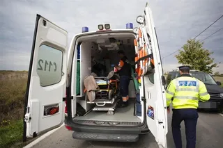 Un polițist în uniformă galbenă și albastră stă lângă spatele unei ambulanțe albe cu ușile deschise pe un drum asfaltat.