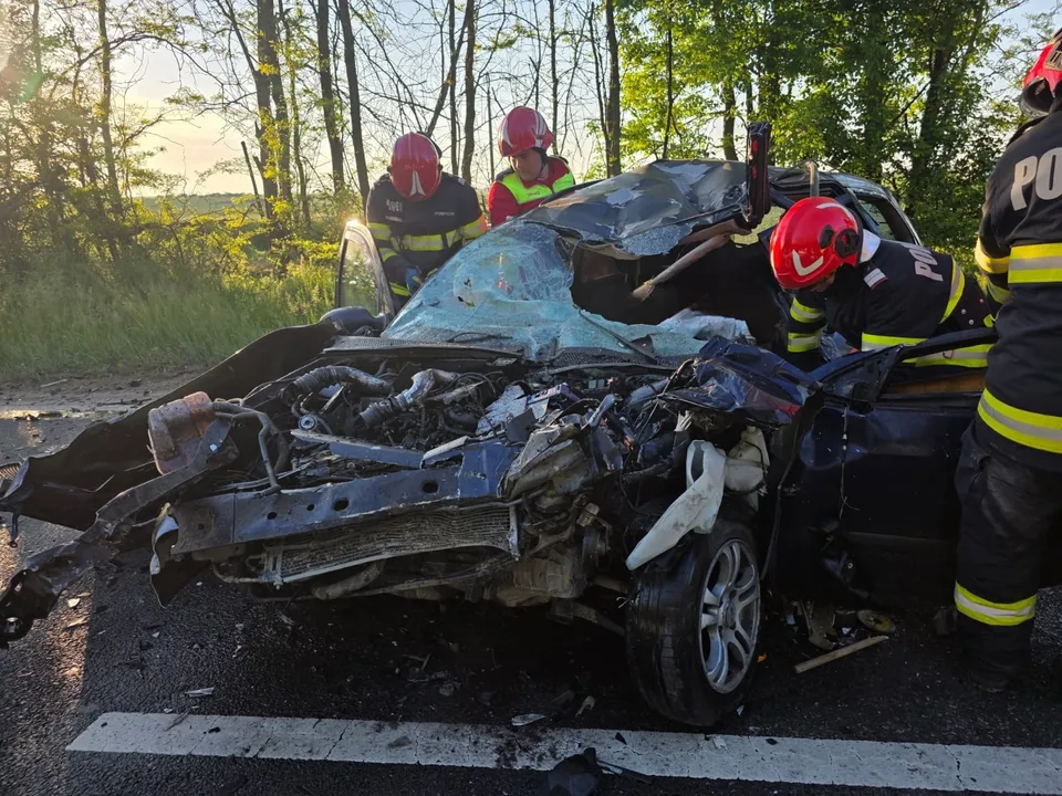 Pompieri cu cască roșie lucrează lângă un autoturism de culoare închisă cu partea frontală puternic distrusă pe un drum asfaltat.