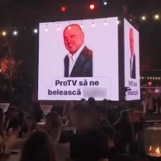 Un ecran mare afișează portretul unui bărbat zâmbitor și un text pe fundal alb, deasupra unei mulțimi de oameni la un eveniment nocturn.