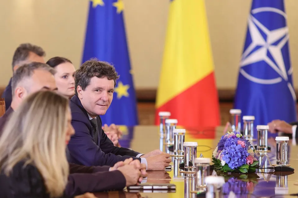 Un bărbat cu părul creț și sacou închis se întoarce spre cameră, stând la o masă de conferință lângă alte persoane și steaguri.
