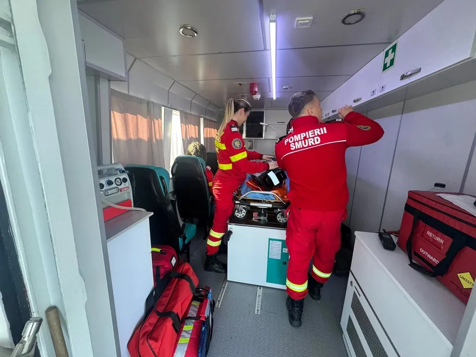Două persoane în uniforme roșii cu inscripții albe se află în interiorul unei ambulanțe, lângă echipamente medicale și saci de transport roșii.
