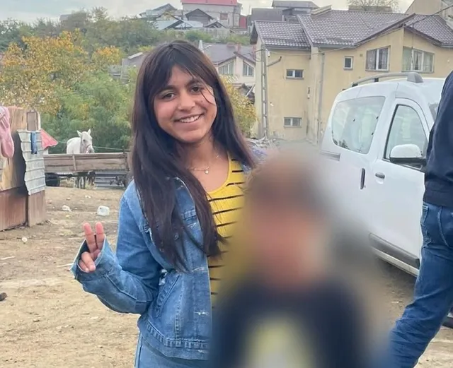 O fată zâmbind poartă o jachetă de denim peste un tricou galben cu dungi negre, stând lângă un vehicul alb.