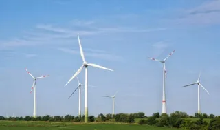 Mai multe turbine eoliene albe sunt amplasate pe un câmp verde sub un cer albastru cu nori subțiri.