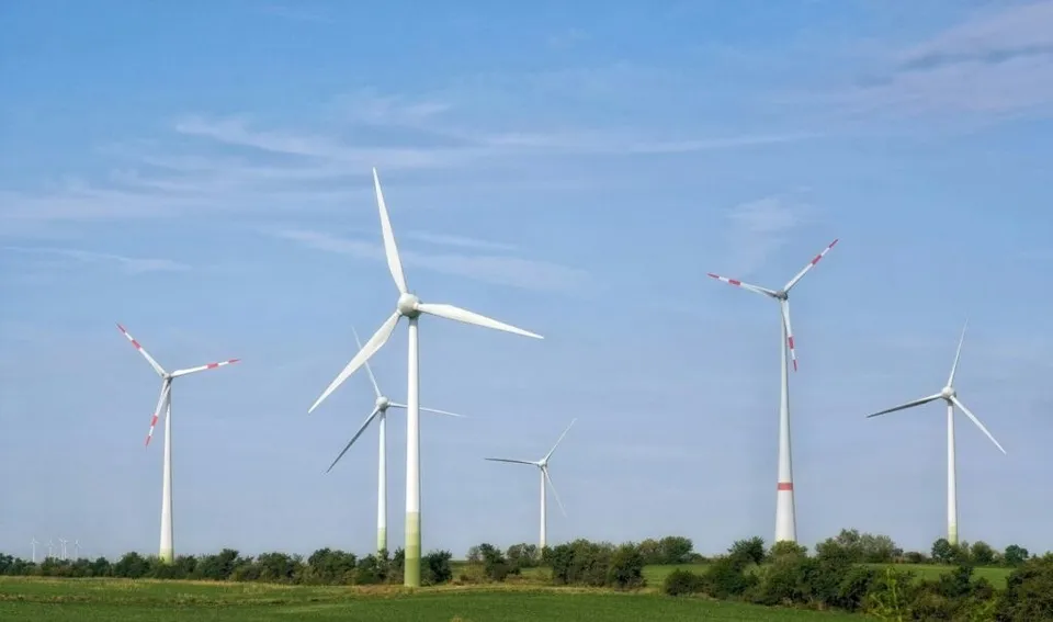 Mai multe turbine eoliene albe sunt amplasate pe un câmp verde sub un cer albastru cu nori subțiri.