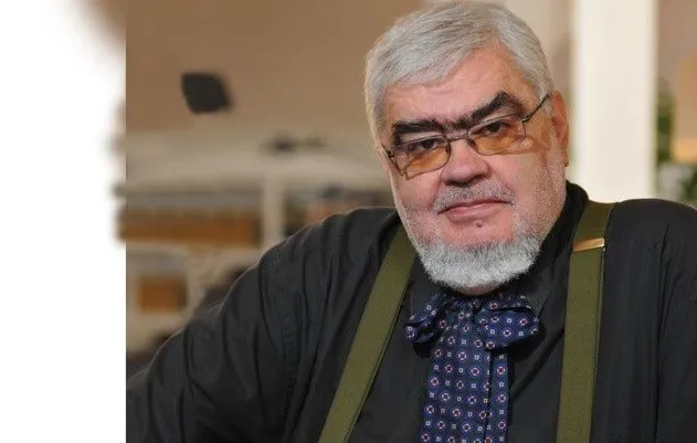 Un bărbat cu părul și barba gri, purtând ochelari, cămașă neagră, bretele verzi și o cravată cu modele geometrice, privește spre cameră.