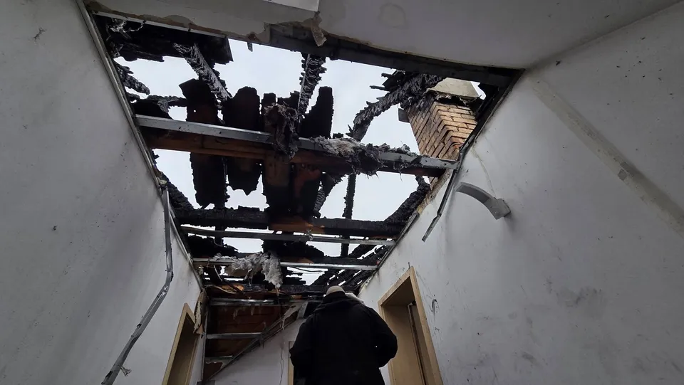 O persoană cu spatele poartă o haină neagră sub un tavan de interior cu o porțiune de structură de lemn arsă și deschisă spre exterior.