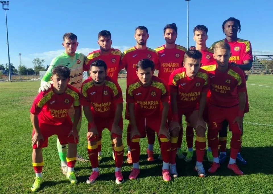 O echipă de fotbaliști în echipamente roșii și galbene posează pentru o fotografie de grup pe un teren de fotbal sub un cer senin.