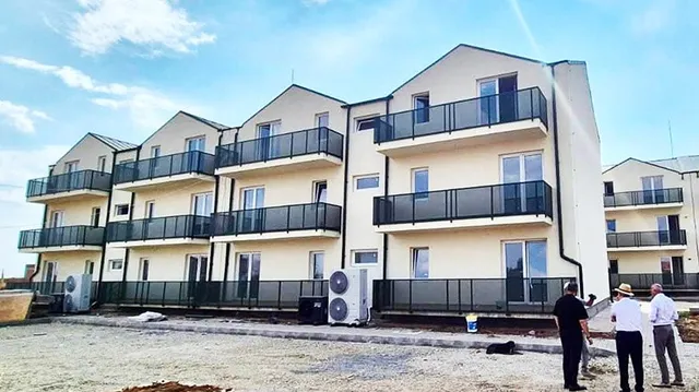 Un grup de trei bărbați stă pe un teren de pietriș în fața unui bloc de apartamente cu balcoane negre și unități de aer condiționat exterioare.