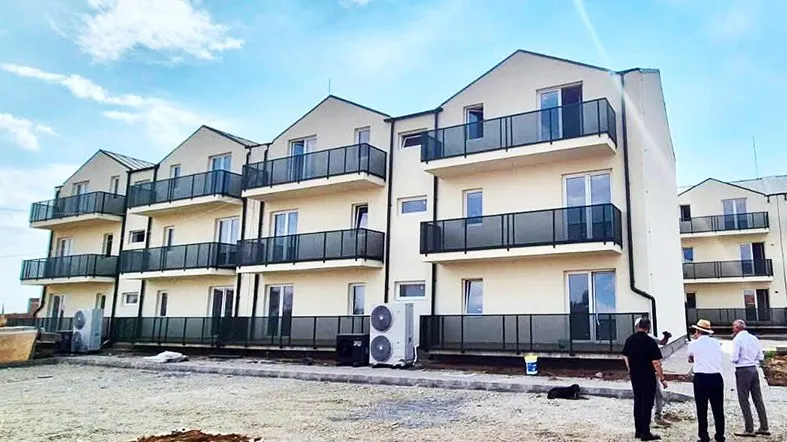 Un grup de trei bărbați stă pe un teren de pietriș în fața unui bloc de apartamente cu balcoane negre și unități de aer condiționat exterioare.