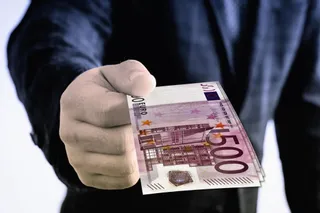 O persoană într-un costum închis oferă un teanc de bancnote de cinci sute de euro către cameră.