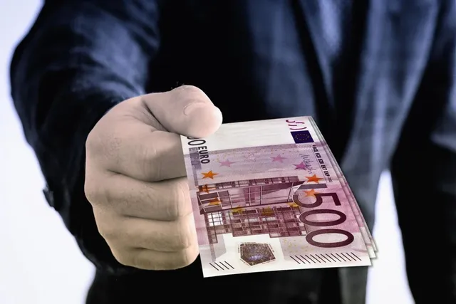 O persoană într-un costum închis oferă un teanc de bancnote de cinci sute de euro către cameră.
