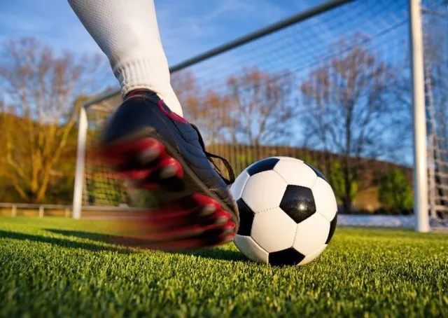 Un picior cu șosetă albă și pantof de fotbal negru lovește un fotbal alb cu pete negre pe un teren de iarbă artificială.