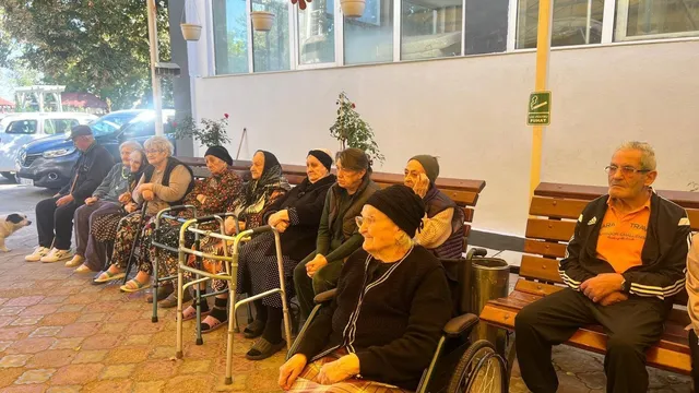 O serie de persoane vârstnice stau așezate pe bănci de lemn și în scaune cu rotile pe o terasă exterioară.