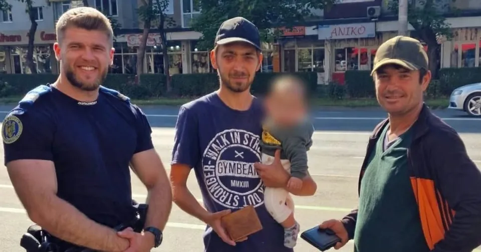 Un jandarm zâmbitor, un bărbat cu un copil în brațe și un alt bărbat, toți trei stând pe o stradă, cu clădiri și mașini în fundal.