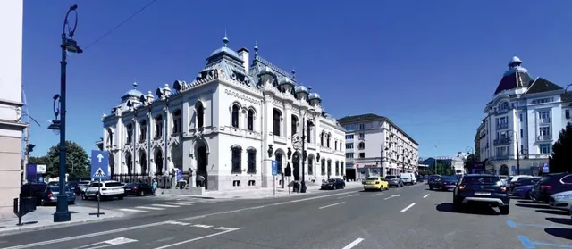 O clădire albă cu arhitectură ornamentată se află lângă o stradă asfaltată cu mașini parcate și în trafic sub un cer senin.
