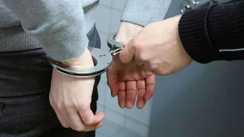 Două mâini sunt prinse în cătușe metalice deasupra unei haine de culoare închisă.