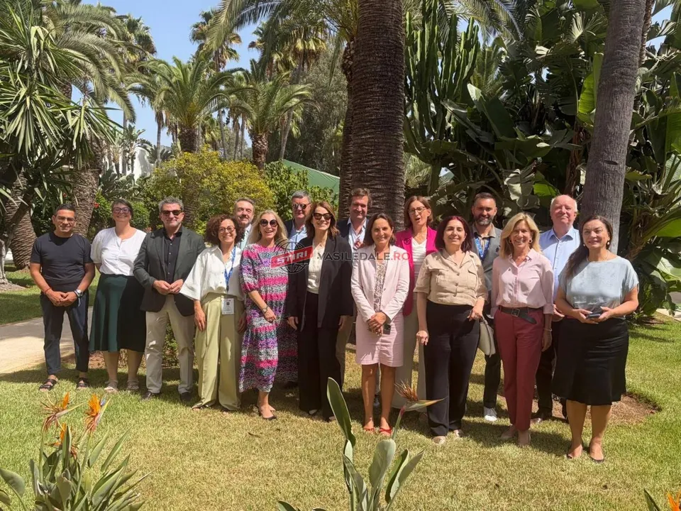 Un grup de persoane stă în rând pe un covor de iarbă verde, în fața unor palmieri și a unor plante verzi sub un cer senin.