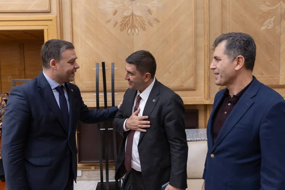Trei bărbați în costume purtând cămași și cravate stau într-o încăpere cu pereți din lemn decorat.