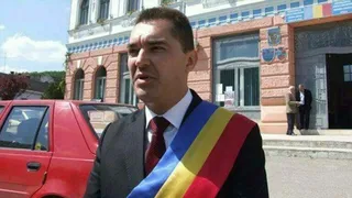Un bărbat îmbrăcat la costum, cu eșarfă tricoloră, stă în fața unei clădiri cu două persoane în ușă și o mașină roșie în stânga.