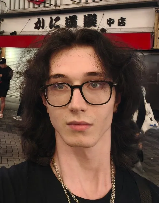 Un bărbat cu părul lung și ochelari cu rame negre poartă un lanț metalic la gât, fiind surprins într-un cadru de tip selfie.