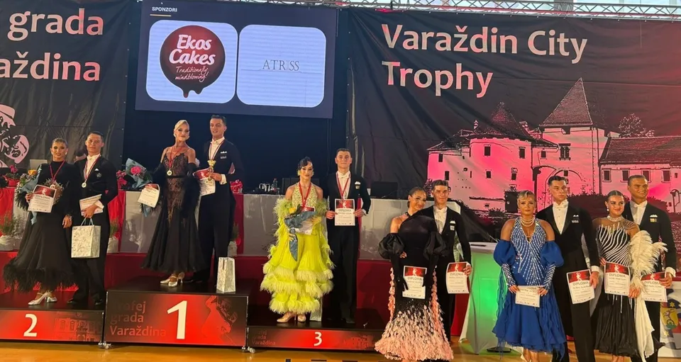 O grupare de dansatori în costume de gala stă pe un podium marcat cu numerele unu, doi și trei în fața unui panou publicitar.