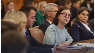 O femeie cu ochelari roșii și bluză deschisă la culoare stă la o masă de conferință lângă alți oameni în interiorul unei încăperi.