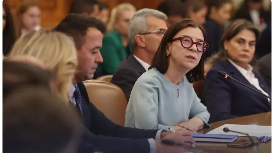 O femeie cu ochelari roșii și bluză deschisă la culoare stă la o masă de conferință lângă alți oameni în interiorul unei încăperi.