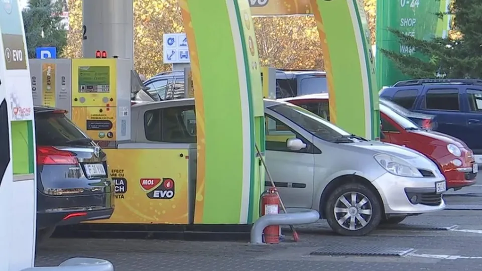 Mașini sunt parcate la o stație de alimentare lângă pompe și panouri de afișaj galbene și verzi.