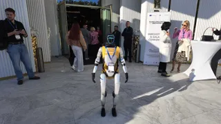 Un robot humanoid cu corp alb și detalii galbene stă în picioare pe o suprafață de marmură lângă un grup de oameni.