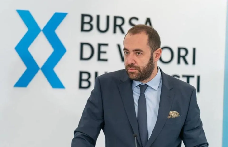 Un bărbat cu barbă, purtând sacou închis la culoare, cămașă albastră și cravată, stă în fața unui perete alb cu un logo și text negru.
