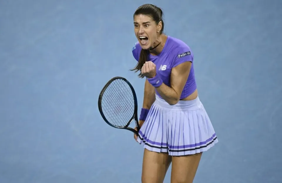 O femeie cu o raketă de tenis în mână poartă o echipă sportivă mov și strigă cu pumnul strâns ridicat pe un teren albastru.