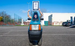 Un bărbat stă în interiorul unui vehicul albastru îngust și ridică un cadru alb deasupra capului pe un teren asfaltat.